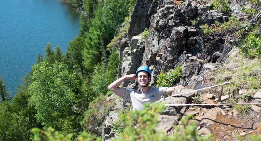 A person wearing safety gear is secured by ropes as they salute the camera while rock climbing, high above a body of water. 